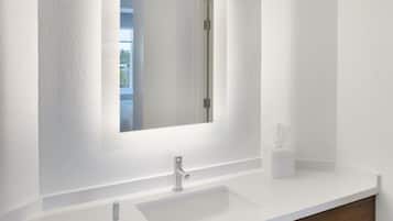 Une salle de bain moderne avec un meuble-lavabo blanc, un grand miroir et une porte visible donnant sur une autre pièce.