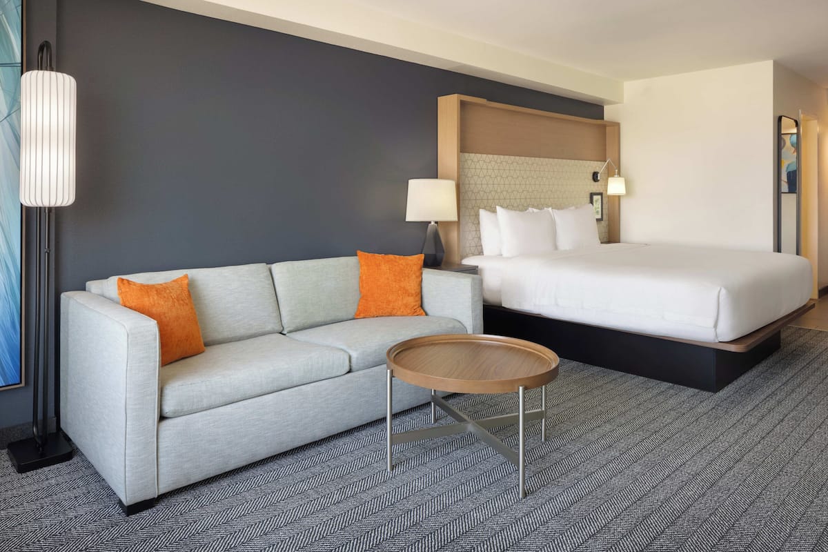 A hotel room with a bed, a sofa, a small round table, a lamp, and a large mirror.