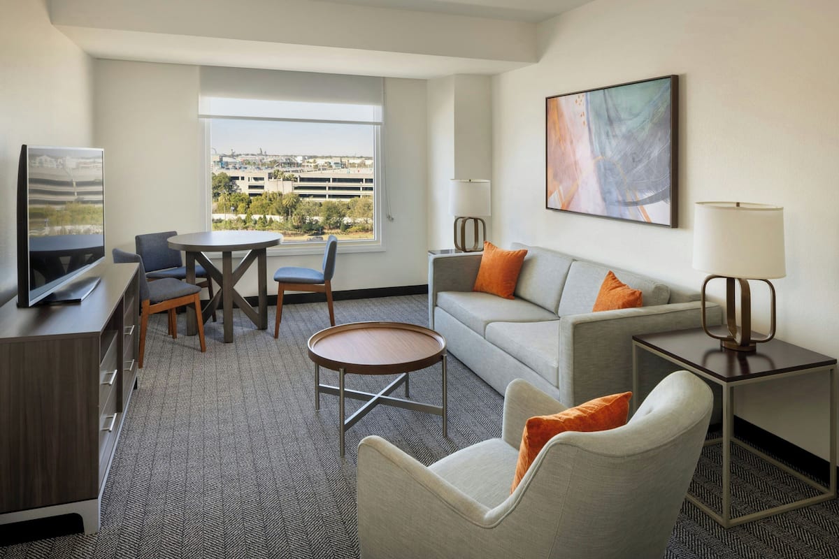 A hotel room with a sofa, armchair, dining table, TV, and a large window with a city view.