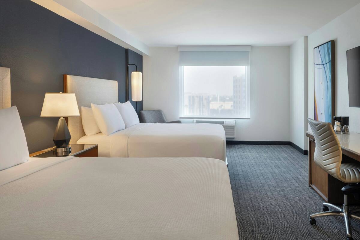 A hotel room with two beds, a desk, a chair, a lamp, a large window, and a city view.