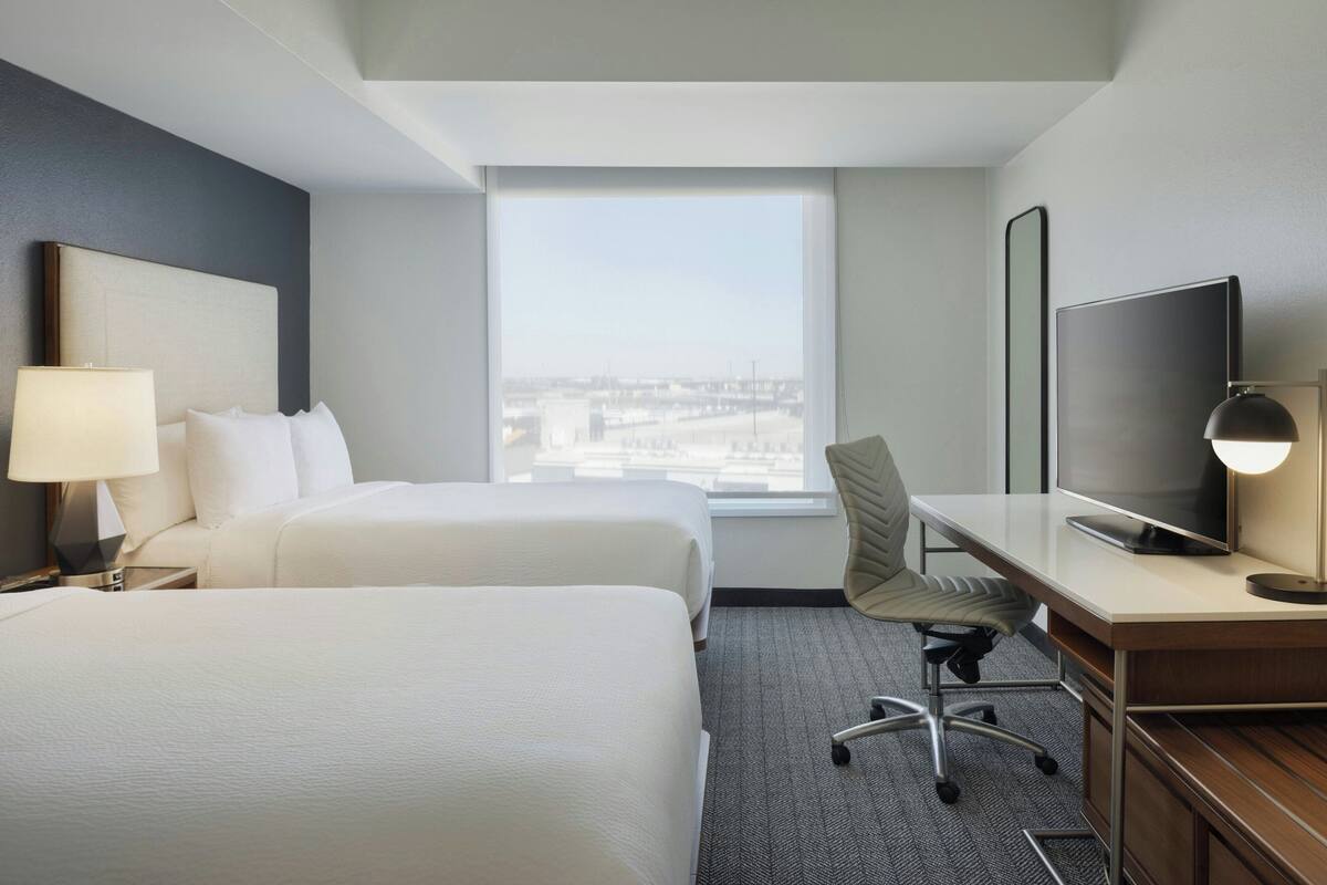 A hotel room with two beds, a desk, a chair, a TV, and a large window with a city view.