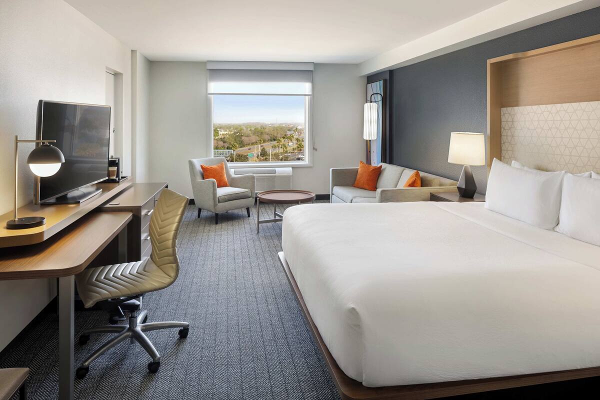 A hotel room with a bed, desk, chair, and a view of the city.
