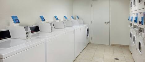 Un cuarto de lavandería con varias lavadoras y secadoras, piso de baldosas y una puerta que da a otra habitación.
