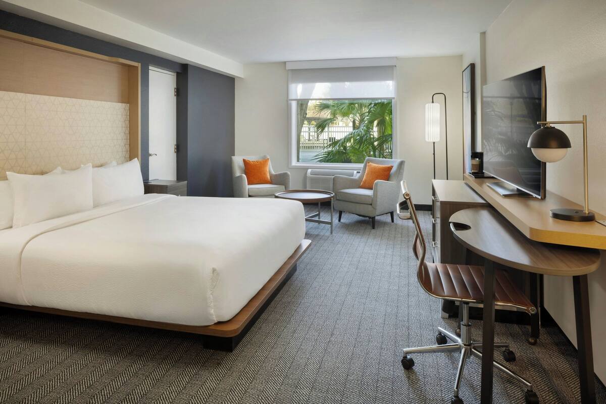 A hotel room with a large bed, a desk with a TV, a chair, and a small table.