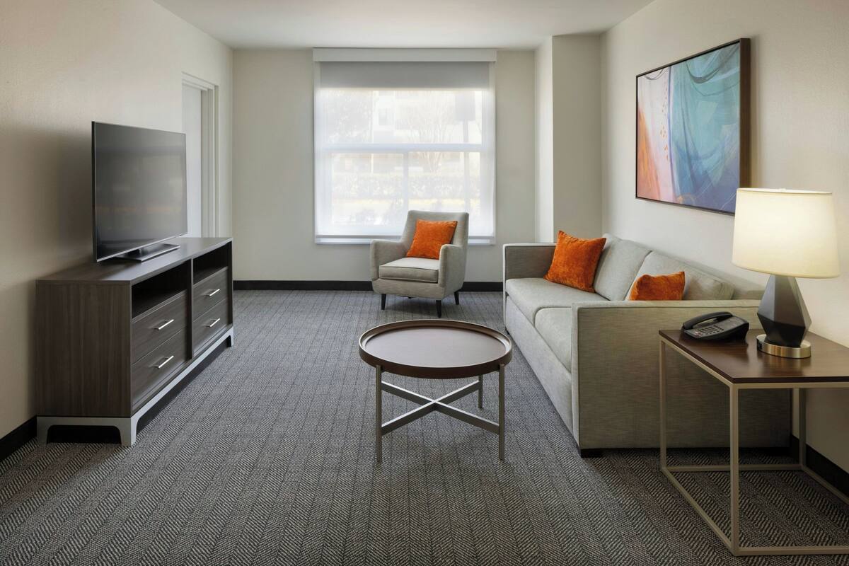 A hotel room with a flat-screen TV, a sofa, a chair, a small table, and a lamp.