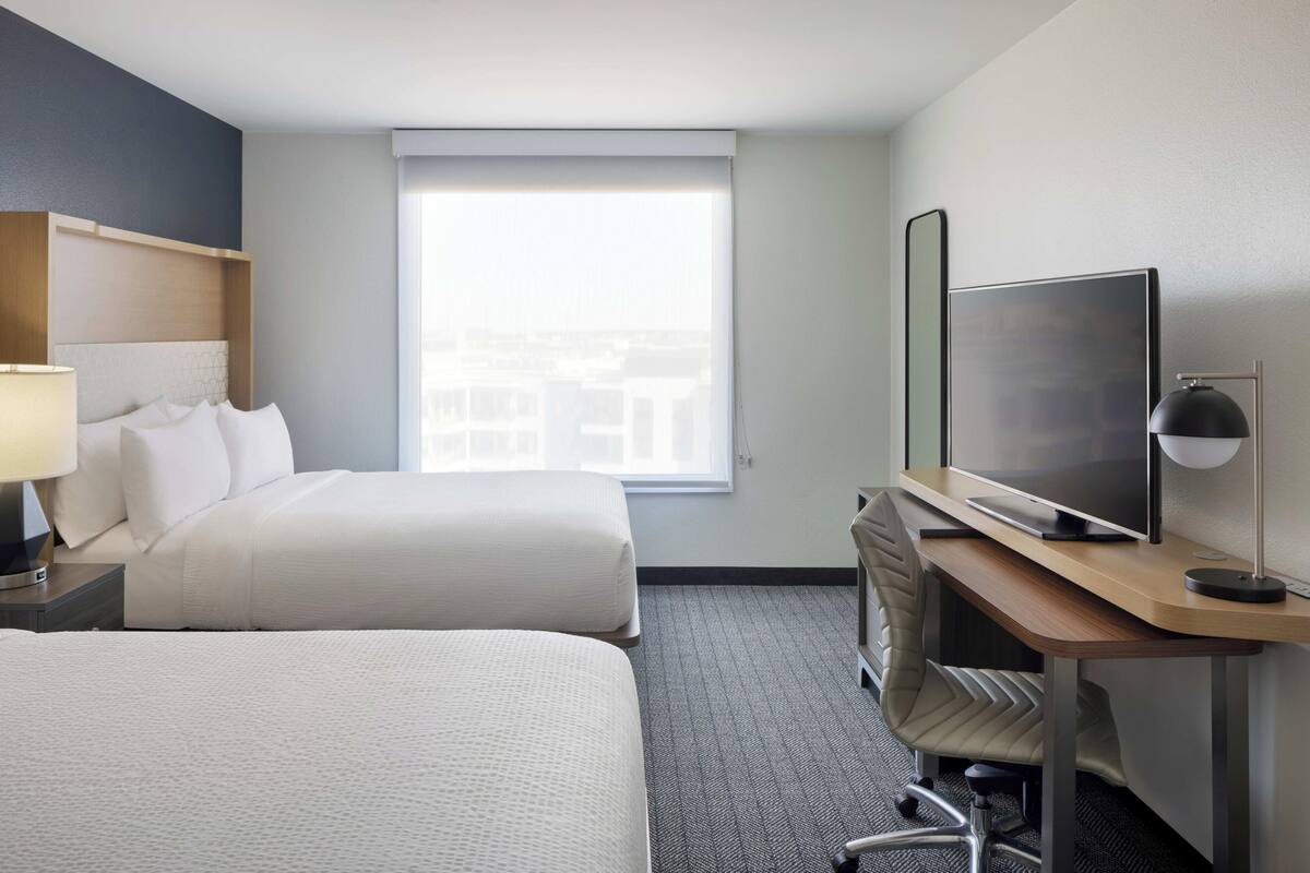 A hotel room with two beds, a desk with a TV, a chair, and a window with a city view.