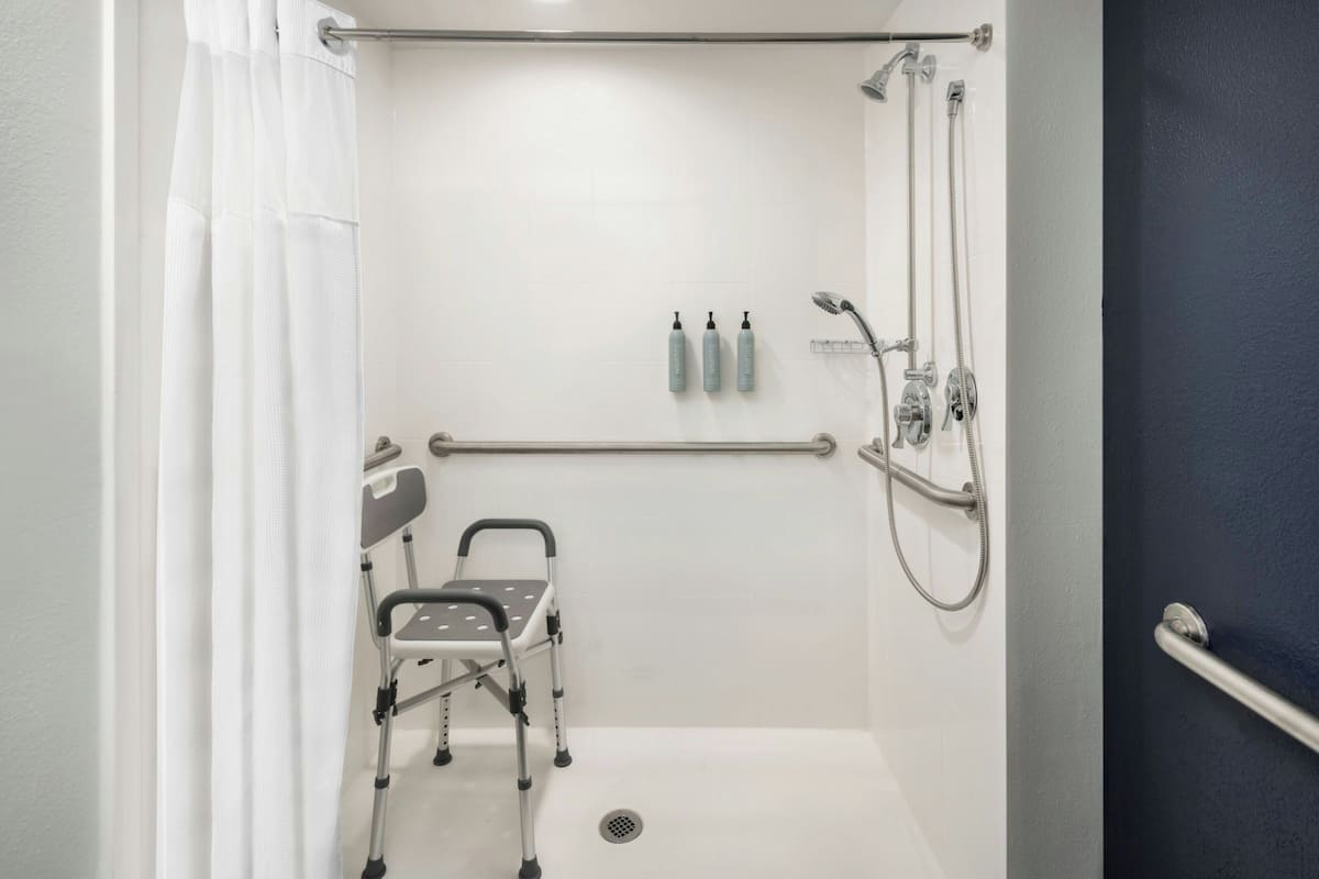 A shower area with a grab bar, a wheelchair, and two bottles of shampoo.