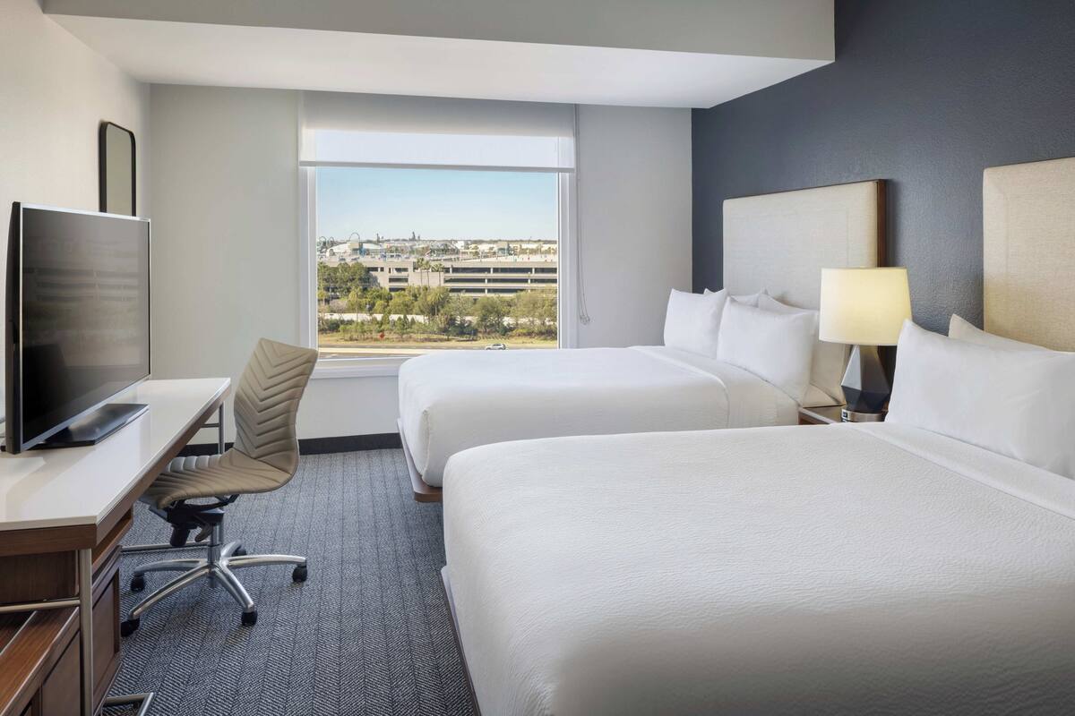 A hotel room with two beds, a desk with a TV, a chair, and a view of a cityscape.