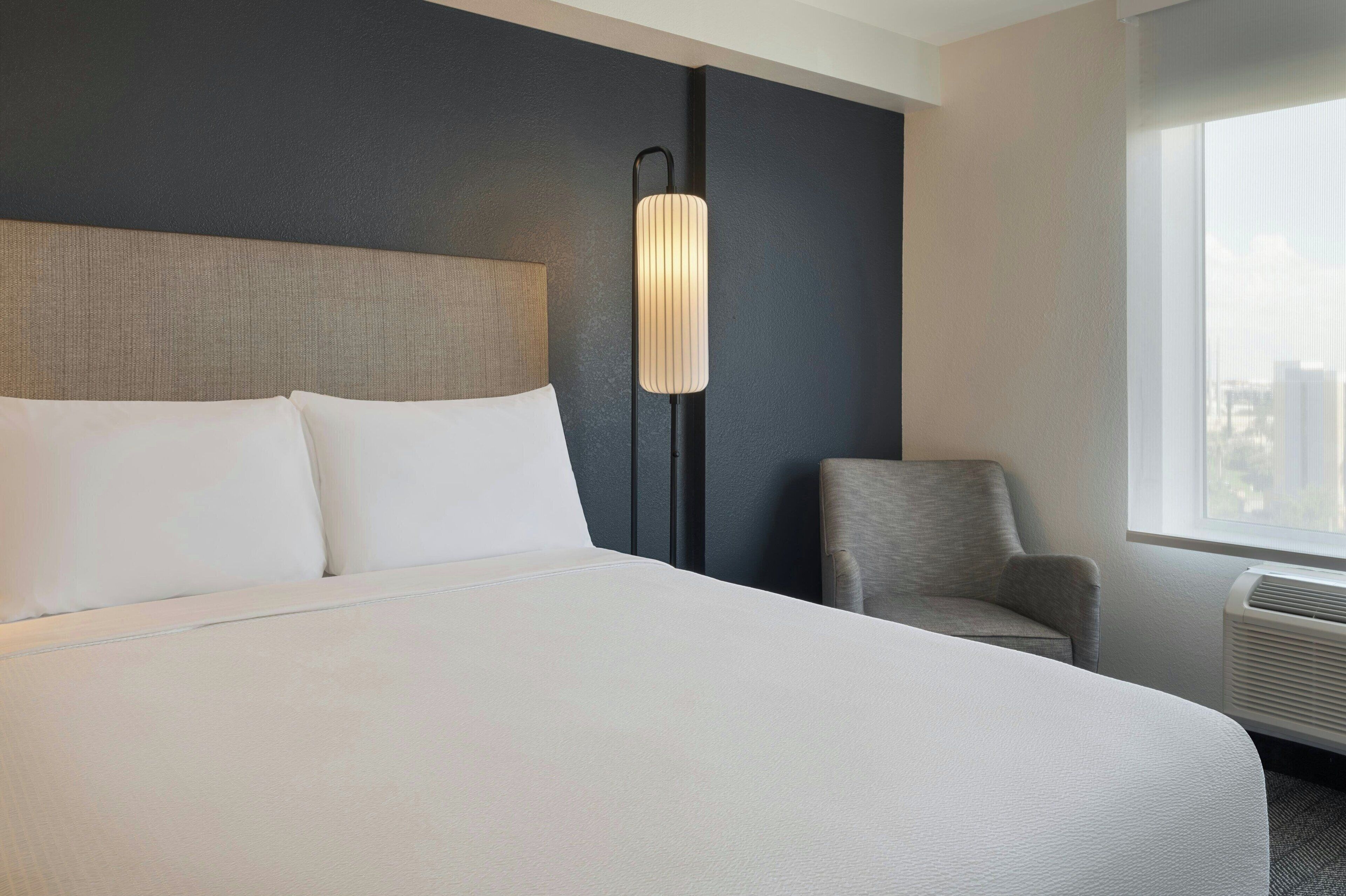 Een hotelkamer met een groot bed, een stoel, een wandlamp en een raam met uitzicht op de stad.