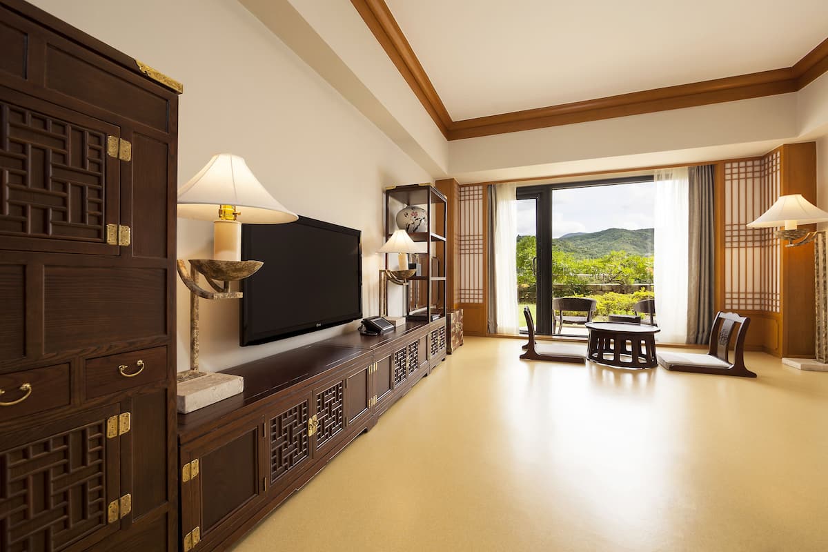 A spacious living room with a large flat-screen TV, wooden cabinets, and a view of greenery through large windows.