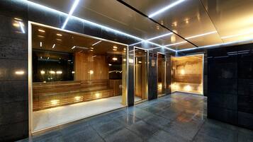 Área de elevador moderna com portas de vidro, um banco de madeira e um painel digital.