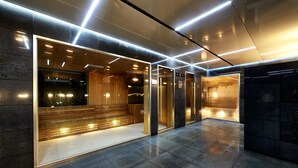 Un área de ascensores moderna con puertas de vidrio, un banco de madera y una pantalla digital.