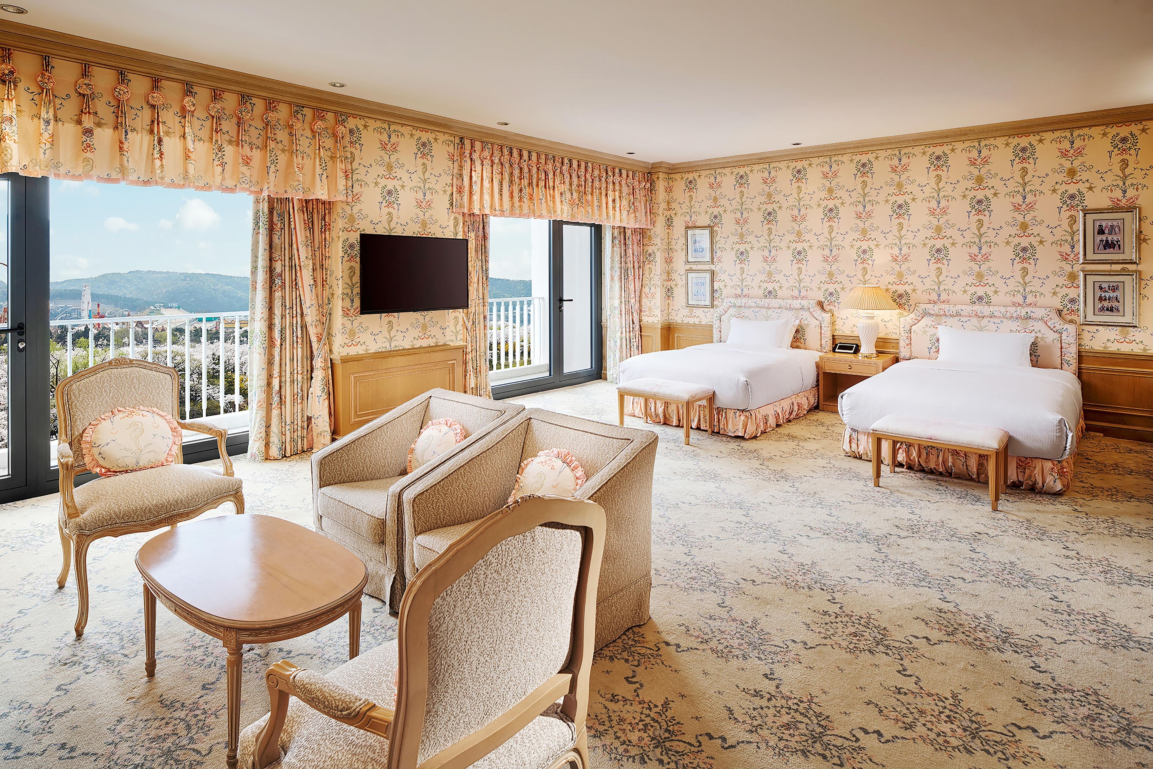 A hotel room with a balcony, a sofa, two beds, a TV, and a desk.