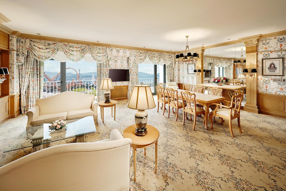 A spacious living room with a sofa, coffee table, dining table, and chairs. There is a TV mounted on the wall, a chandelier, and a balcony with a view of the city.