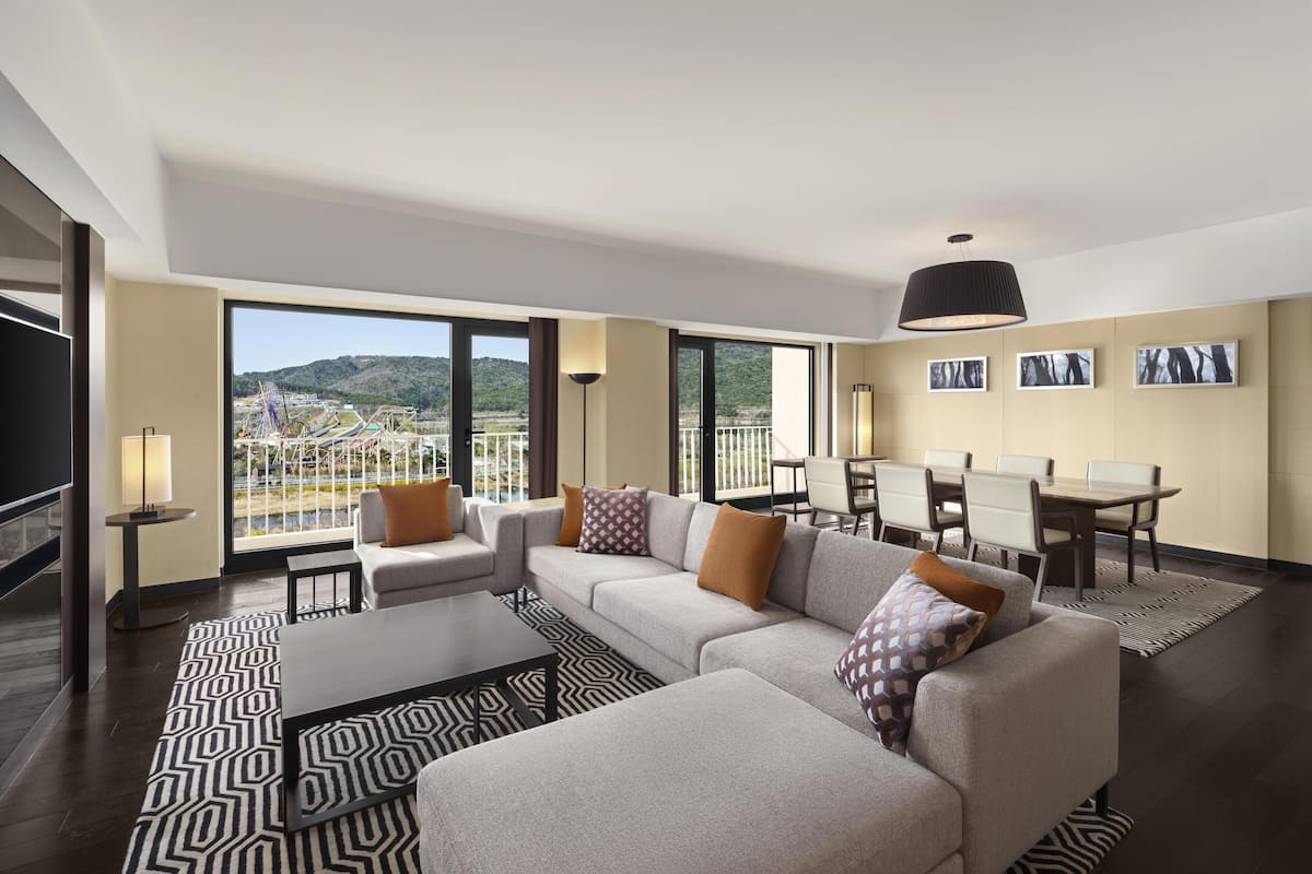 A modern hotel room with a large sofa, a dining table, and a balcony with a view of a park and distant buildings.