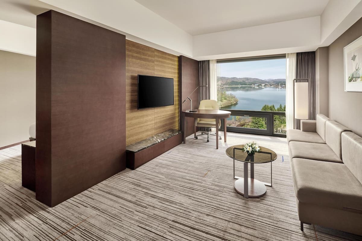 A modern hotel room with a large window, a flat-screen TV, a sofa, a round coffee table, and a small desk with a chair.