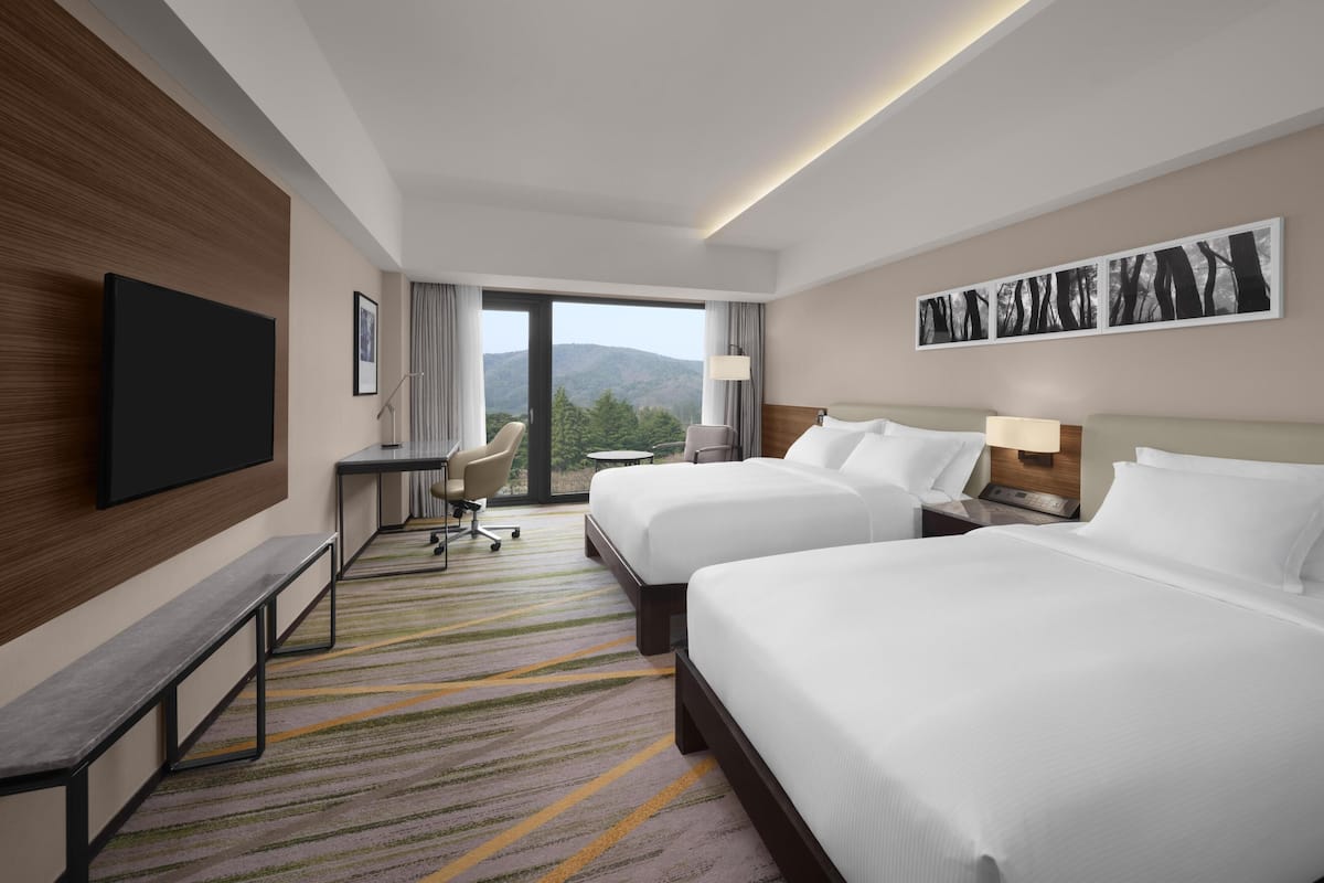 A hotel room with two beds, a desk, a chair, a TV, and a large window with a view of the mountains.