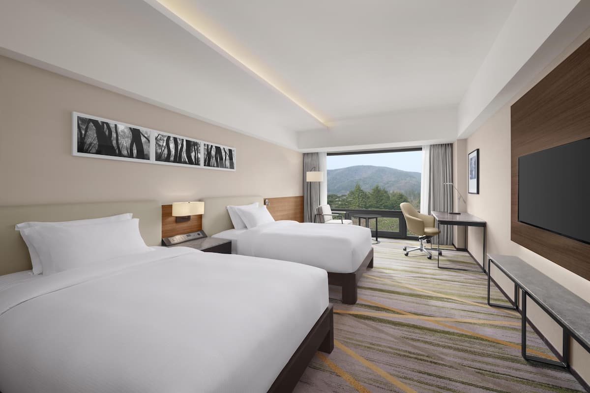A hotel room with two beds, a desk, a chair, a TV, and a balcony with a mountain view.