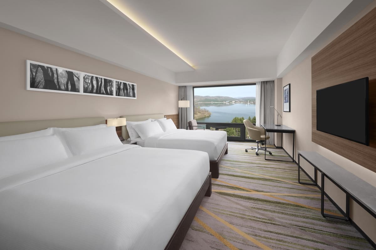 A hotel room with two beds, a TV, a desk, and a view of a lake.