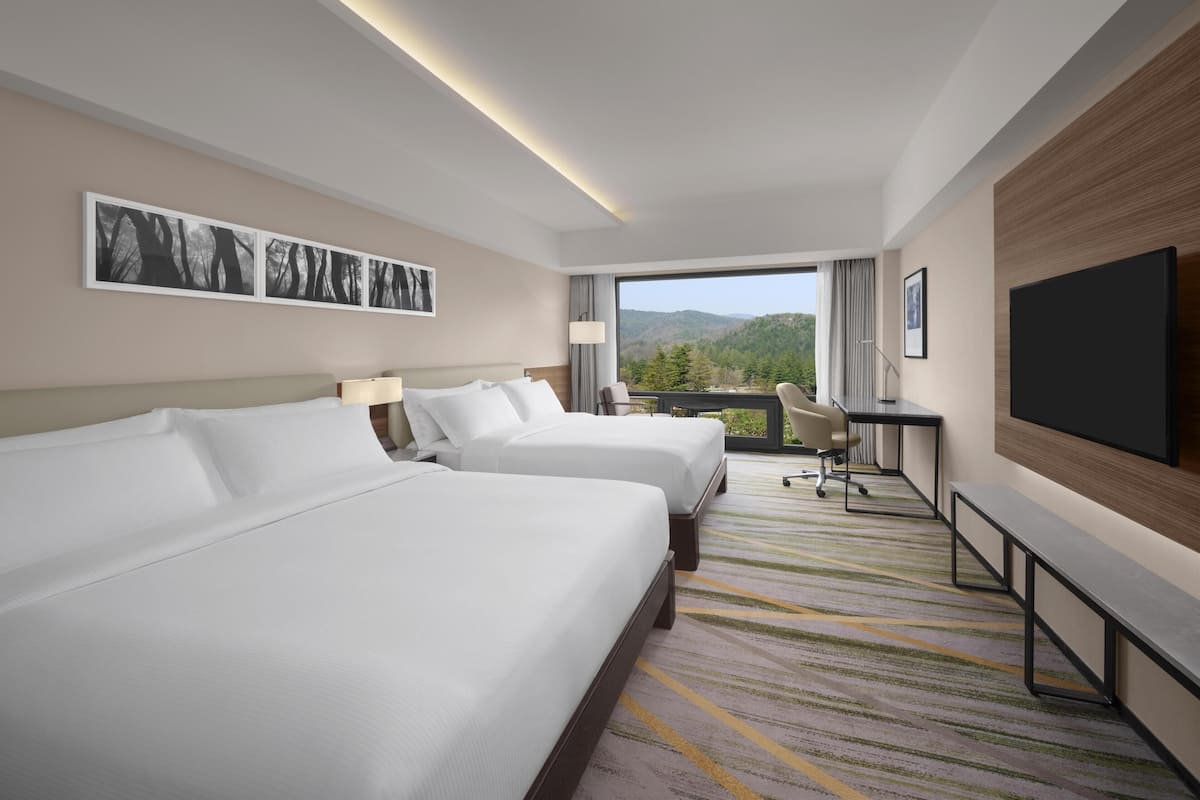 A hotel room with two beds, a desk, a chair, a TV, and a view of the outdoors.