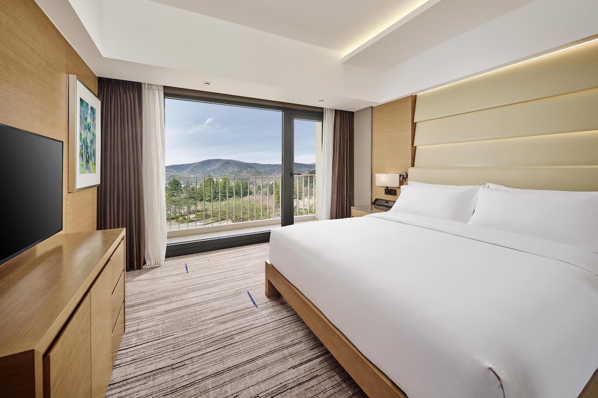 A hotel room with a large bed, a TV, a desk, and a window with a view of mountains.