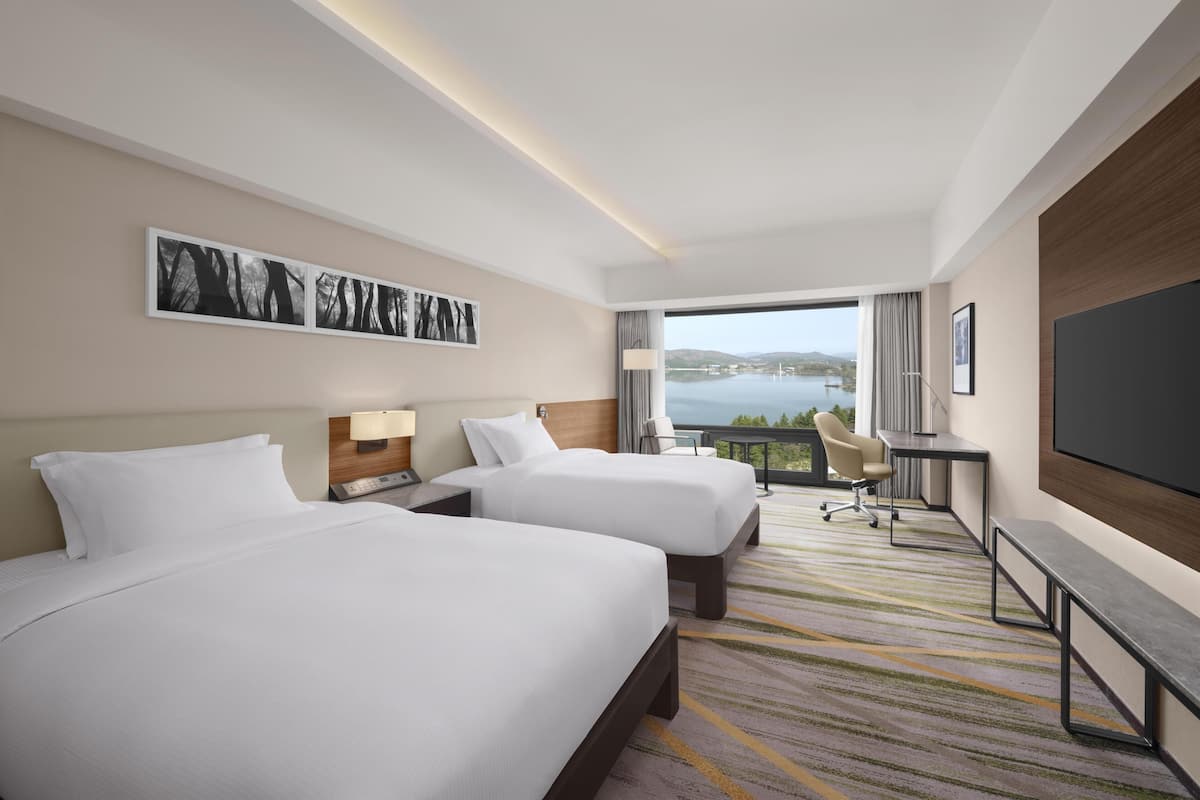 A hotel room with two beds, a desk, a chair, a TV, and a large window with a view of a body of water.