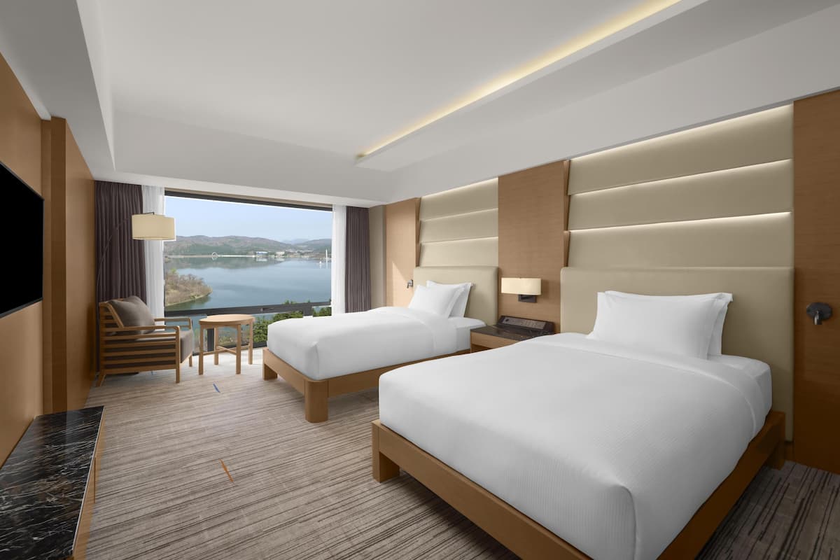 A hotel room with two beds, a TV, a small table, and a view of a lake.