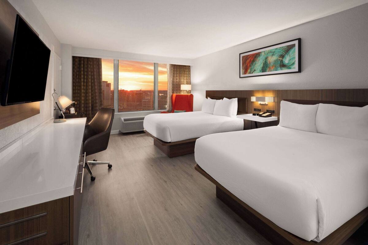 Habitación de hotel con dos camas, un escritorio, una silla, un televisor y un ventanal con vistas a la ciudad.