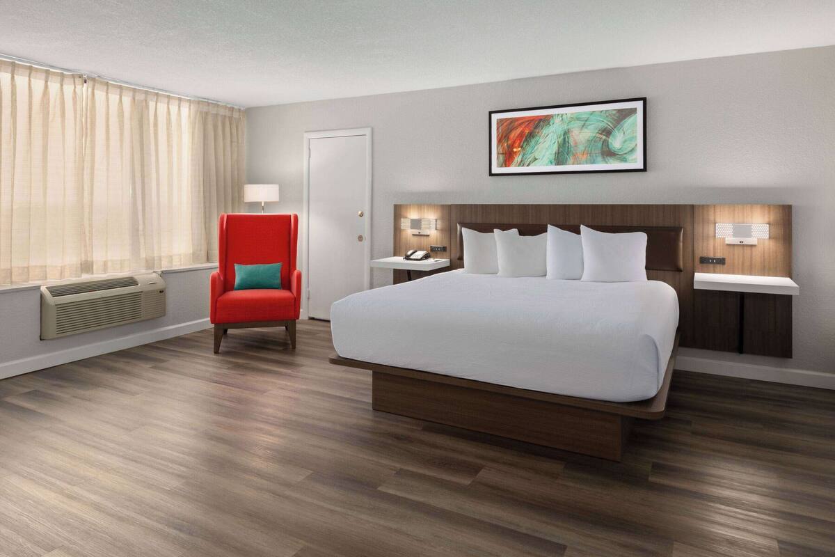 Una habitación de hotel con una cama grande, un sillón rojo, una mesita de noche con lámpara y una ventana con cortinas.