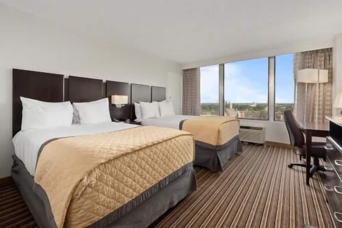 Habitación de hotel con dos camas, un escritorio, una silla y un amplio ventanal con vista a la ciudad.