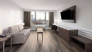 Moderni hotellihuone, jossa on sohva, työpöytä, taulutelevisio ja suuri ikkuna, josta avautuu näkymä kaupunkiin.
