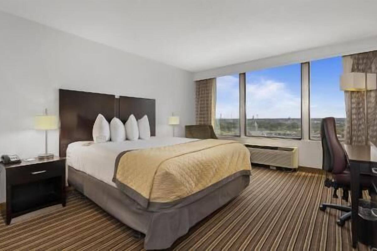 Habitación de hotel con cama, mesitas de noche, escritorio con silla y ventanas amplias con vistas.
