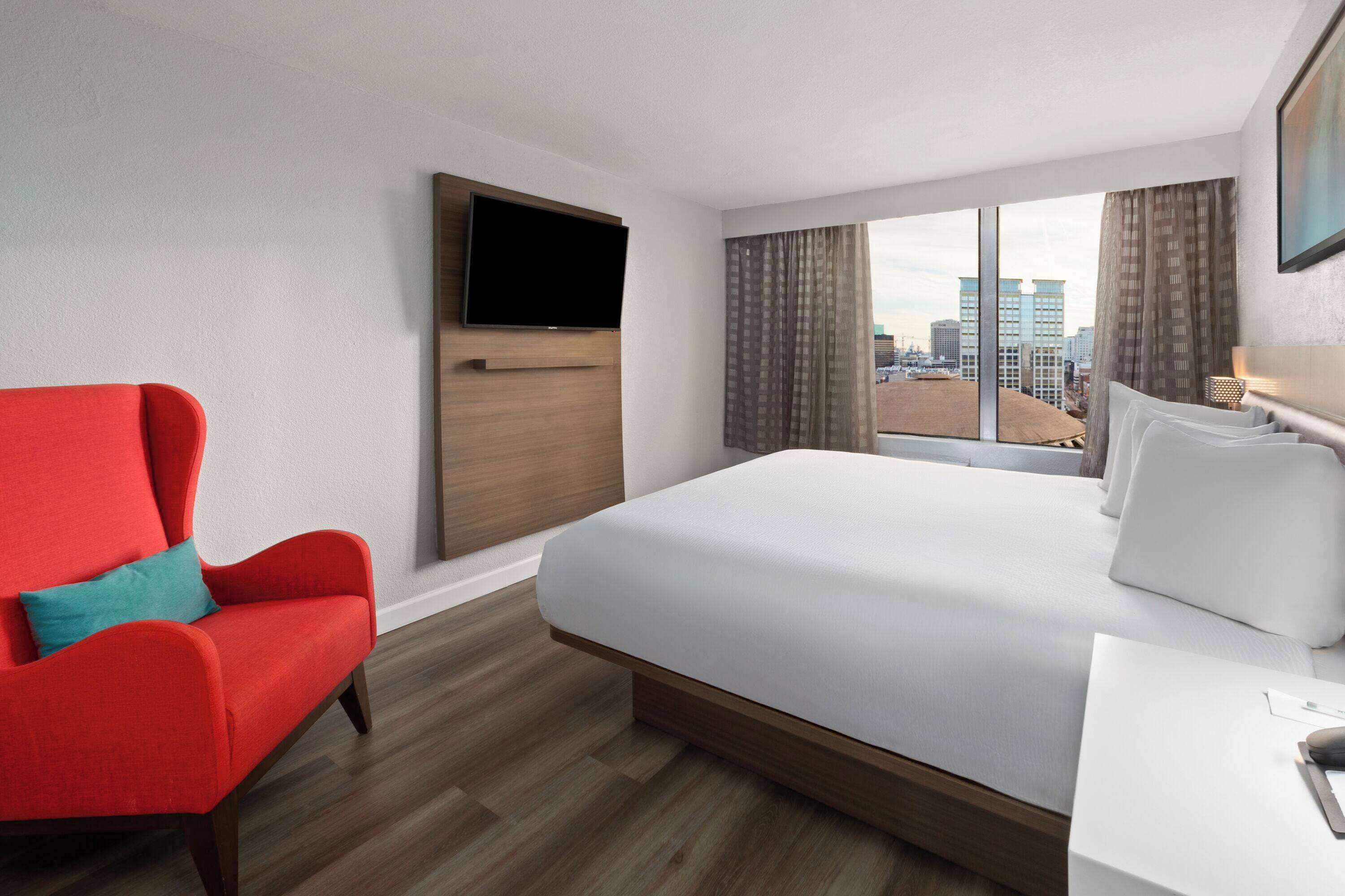 Habitación de hotel con una cama grande, un sillón rojo, un televisor en la pared y vistas a la ciudad desde la ventana.