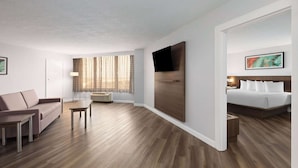 Moderni hotellihuone, jossa on puulattia, sohva, pieni pöytä, valkoisilla lakanoilla varustettu sänky, seinälle asennettu televisio ja ikkuna, jossa on verhot.