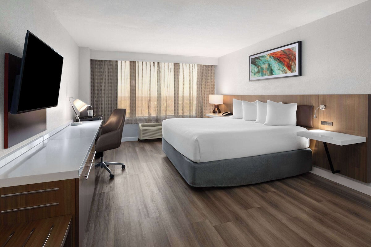 Una habitación de hotel moderna con una cama grande, un escritorio con televisor de pantalla plana, una silla y un ventanal con cortinas.