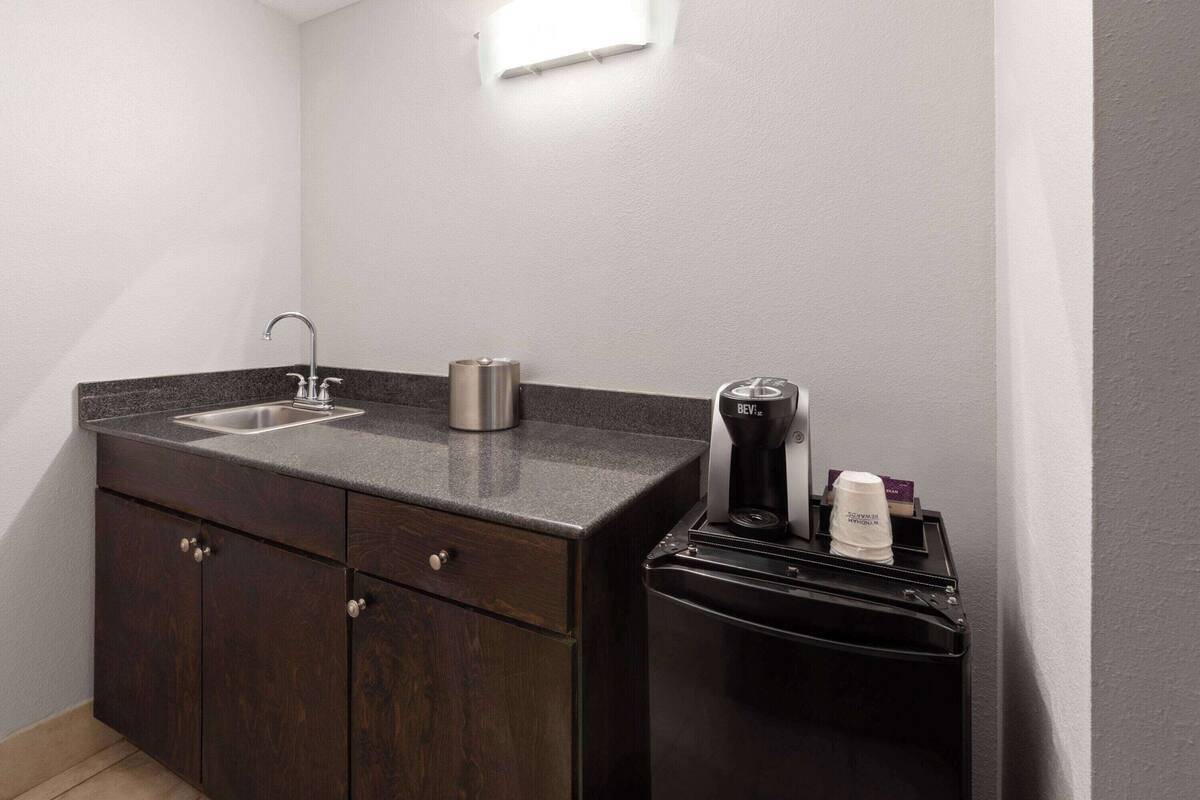 Una cocina compacta con un armario de madera oscura, un lavamanos de acero inoxidable, una cafetera y un refrigerador negro.