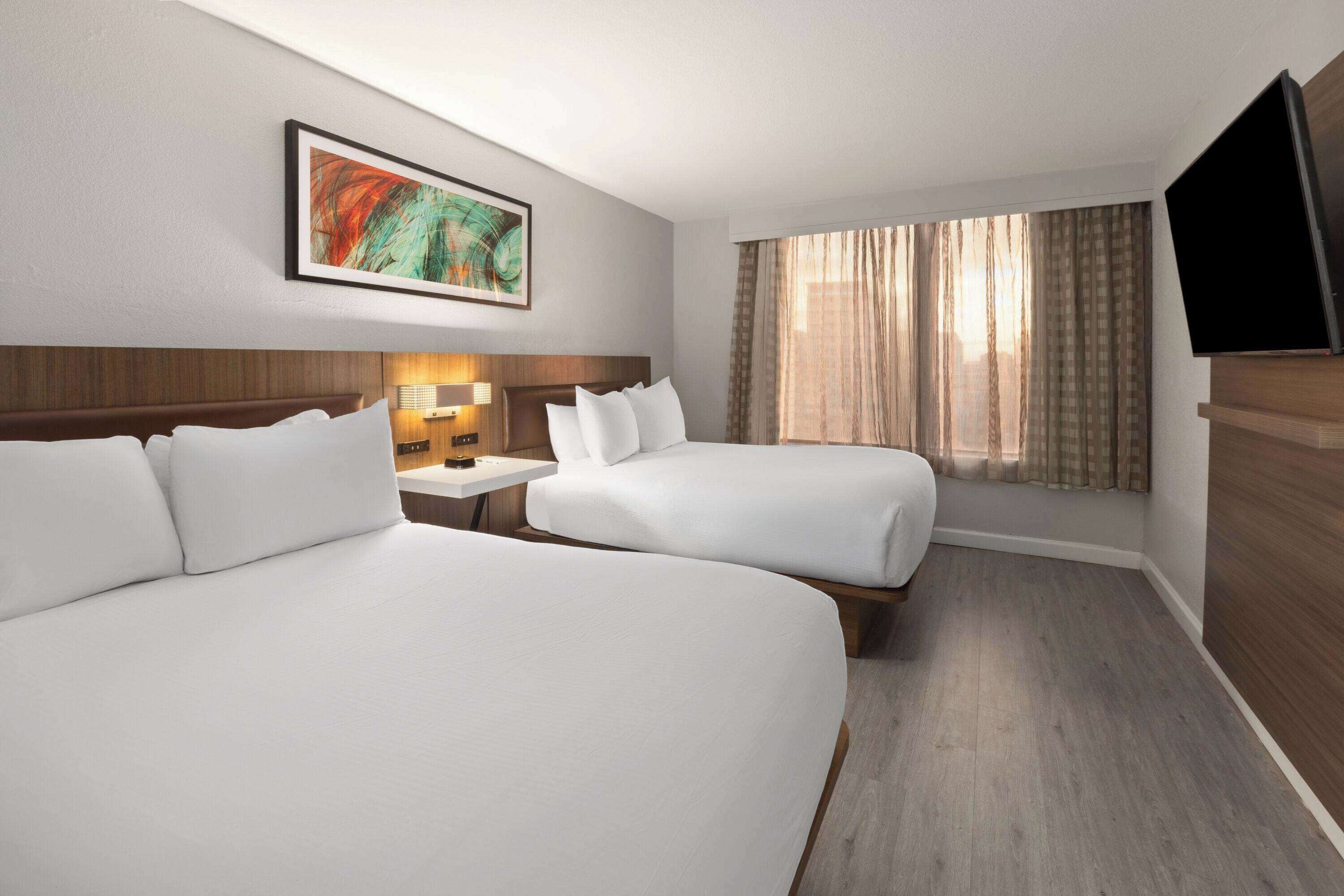 A hotel room with two beds, a flat-screen TV, a painting on the wall, and a window with curtains.