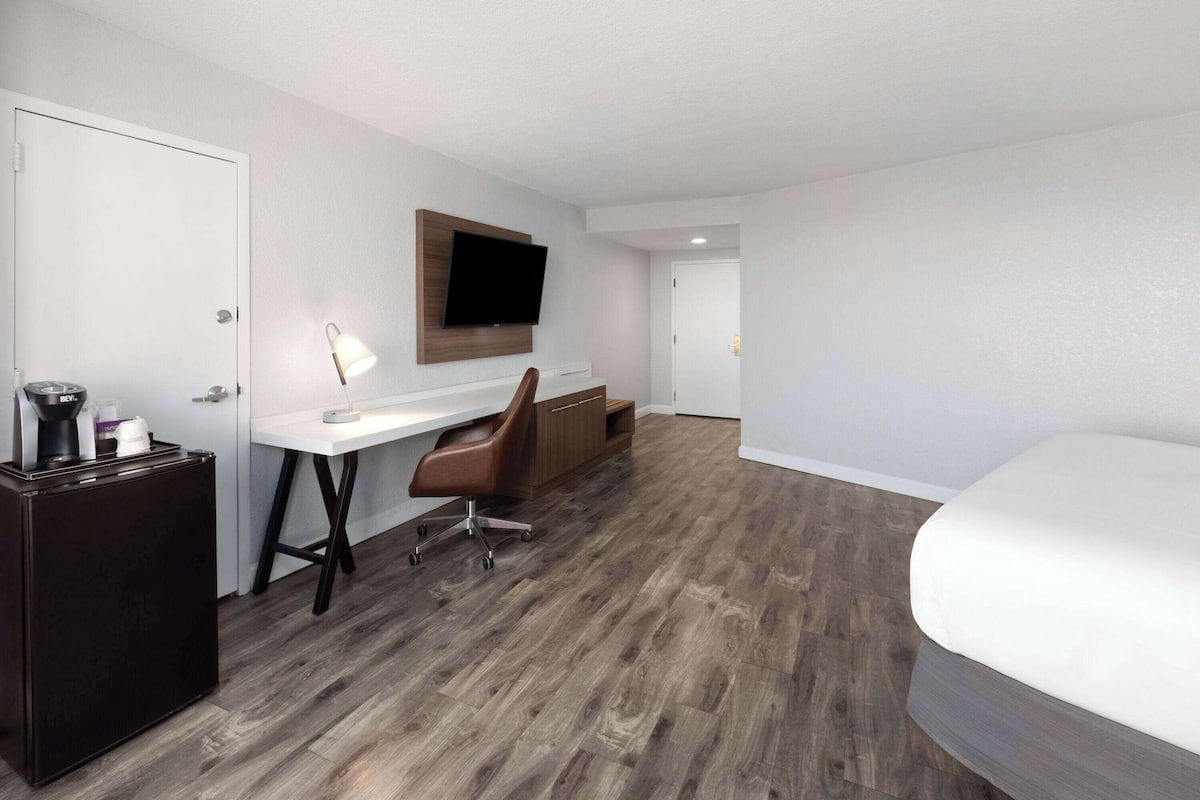 Una habitación de hotel con cama, un escritorio con silla, un televisor montado en la pared y un refrigerador pequeño.