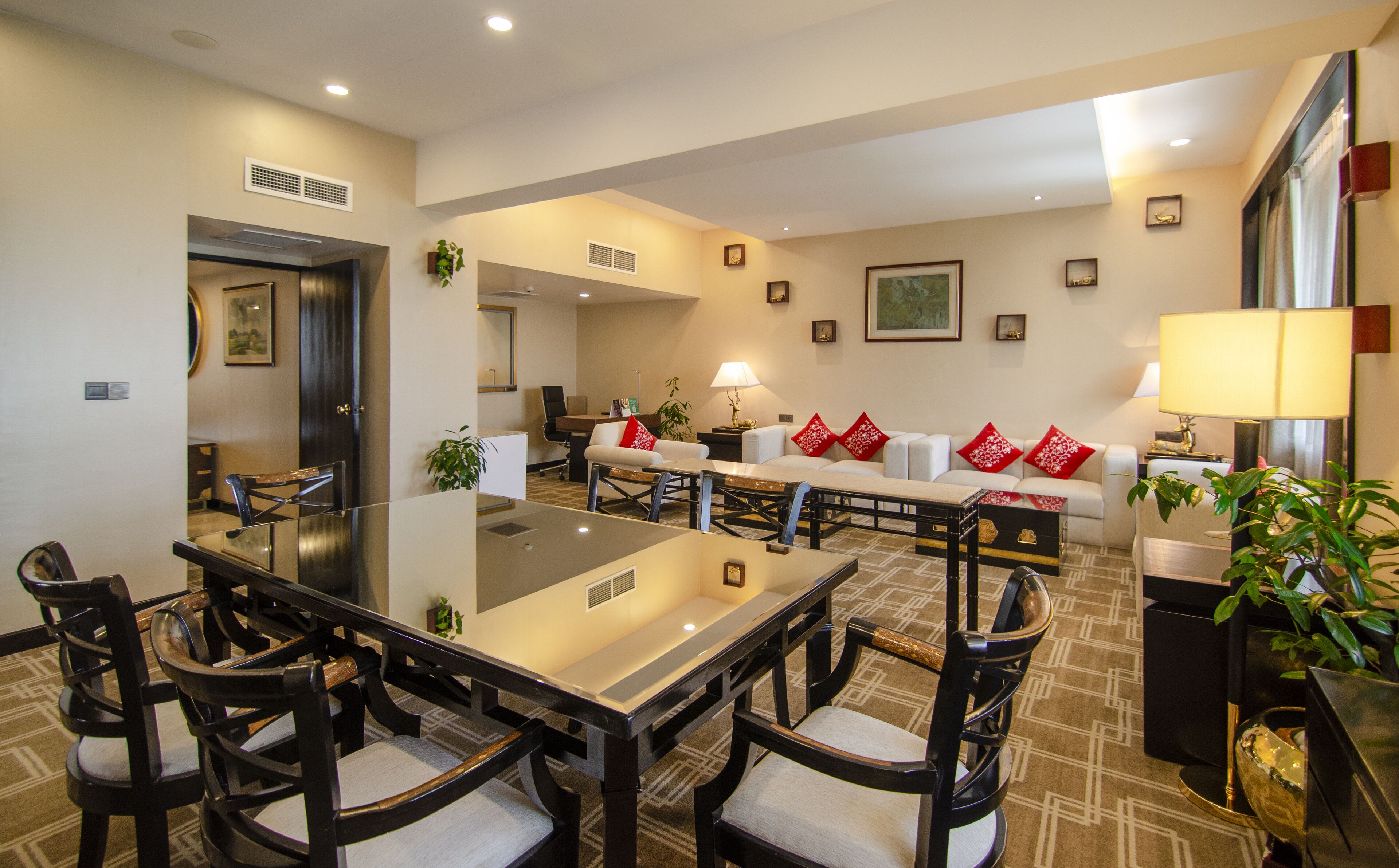 A modern hotel dining area with a glass-top table, dark wooden chairs, a sofa with red cushions, and a lamp.