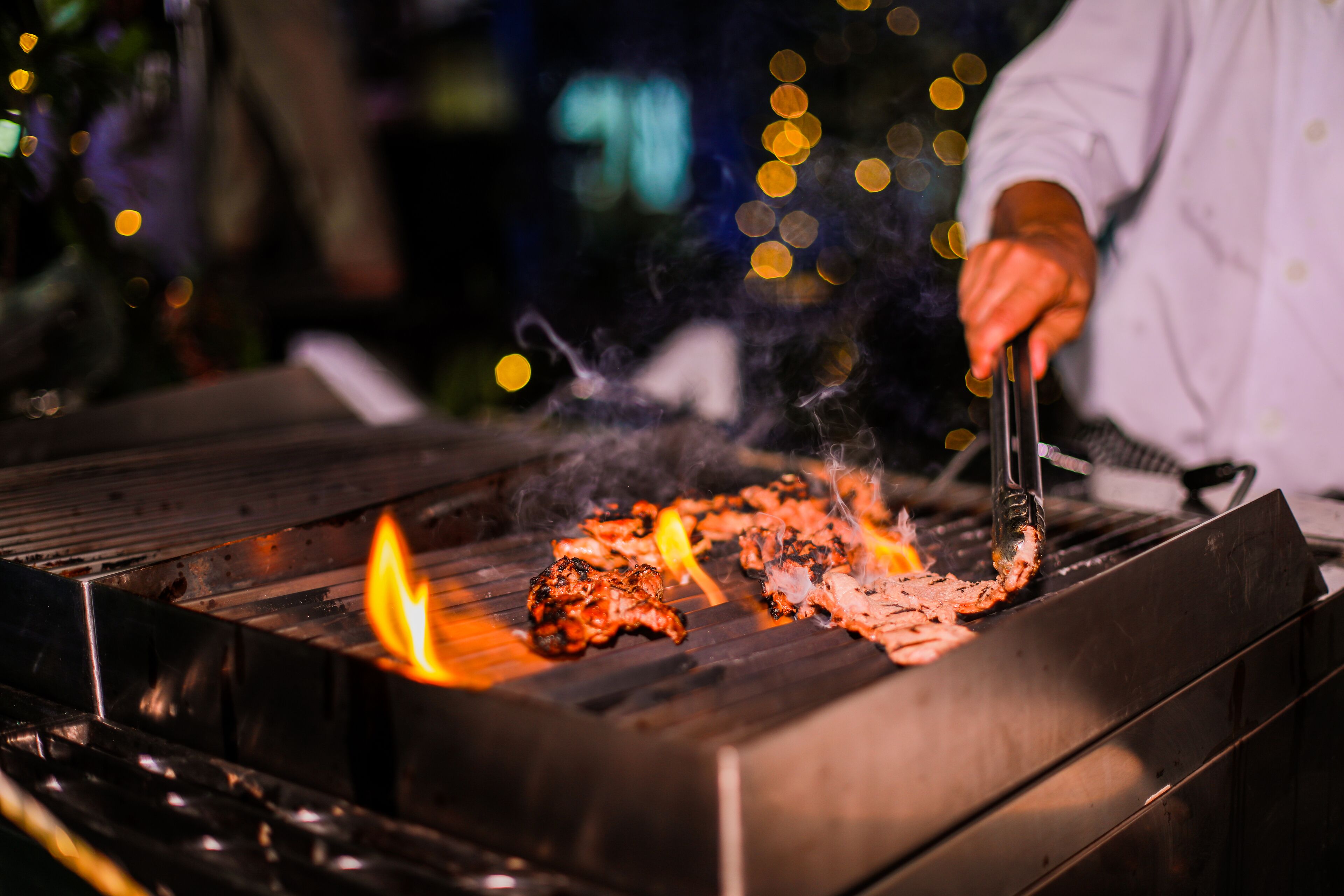 Una parrilla con carne cocinándose, una persona usando pinzas, y un fondo borroso con luces.