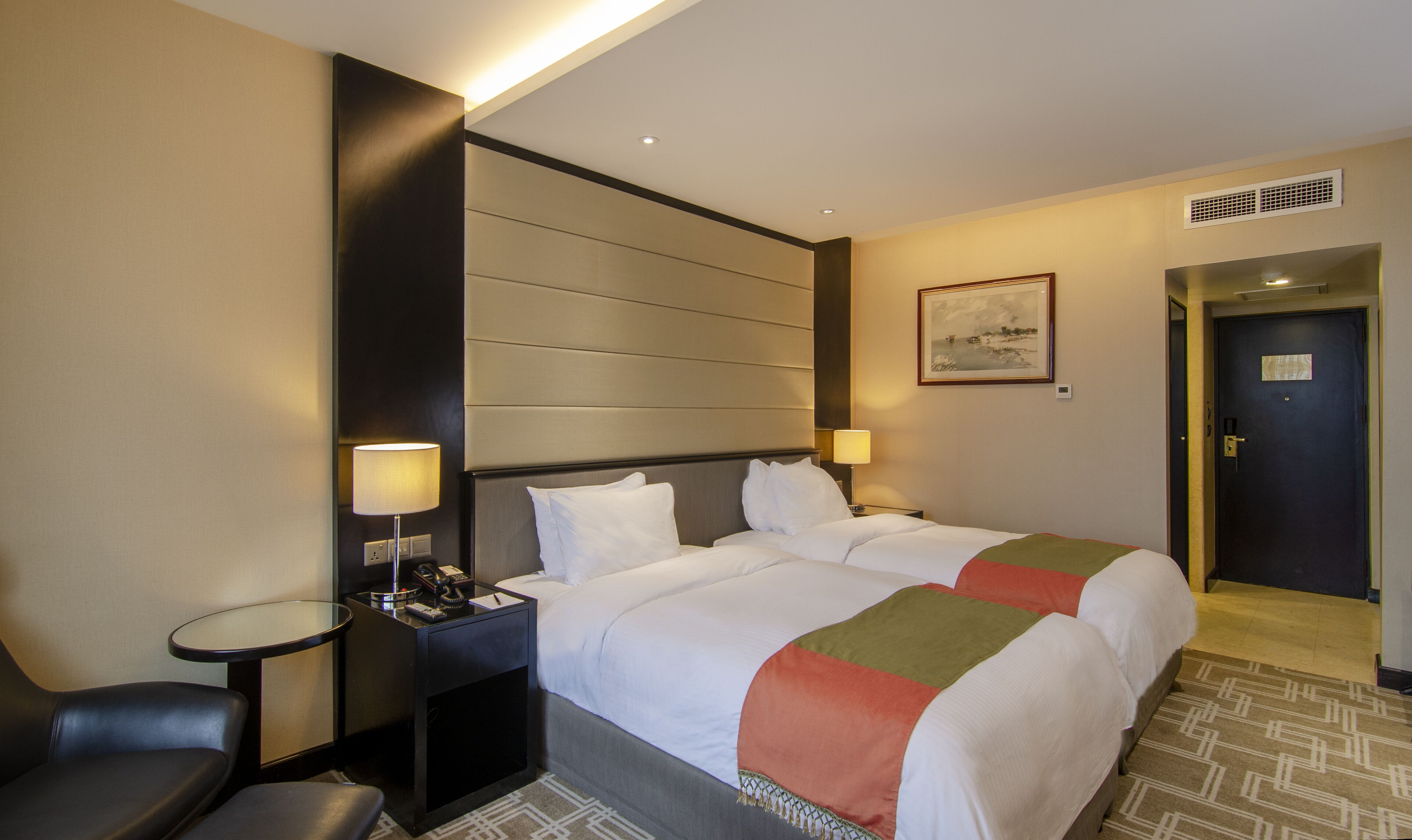 A hotel room with two beds, a desk, a chair, a painting, and a door.