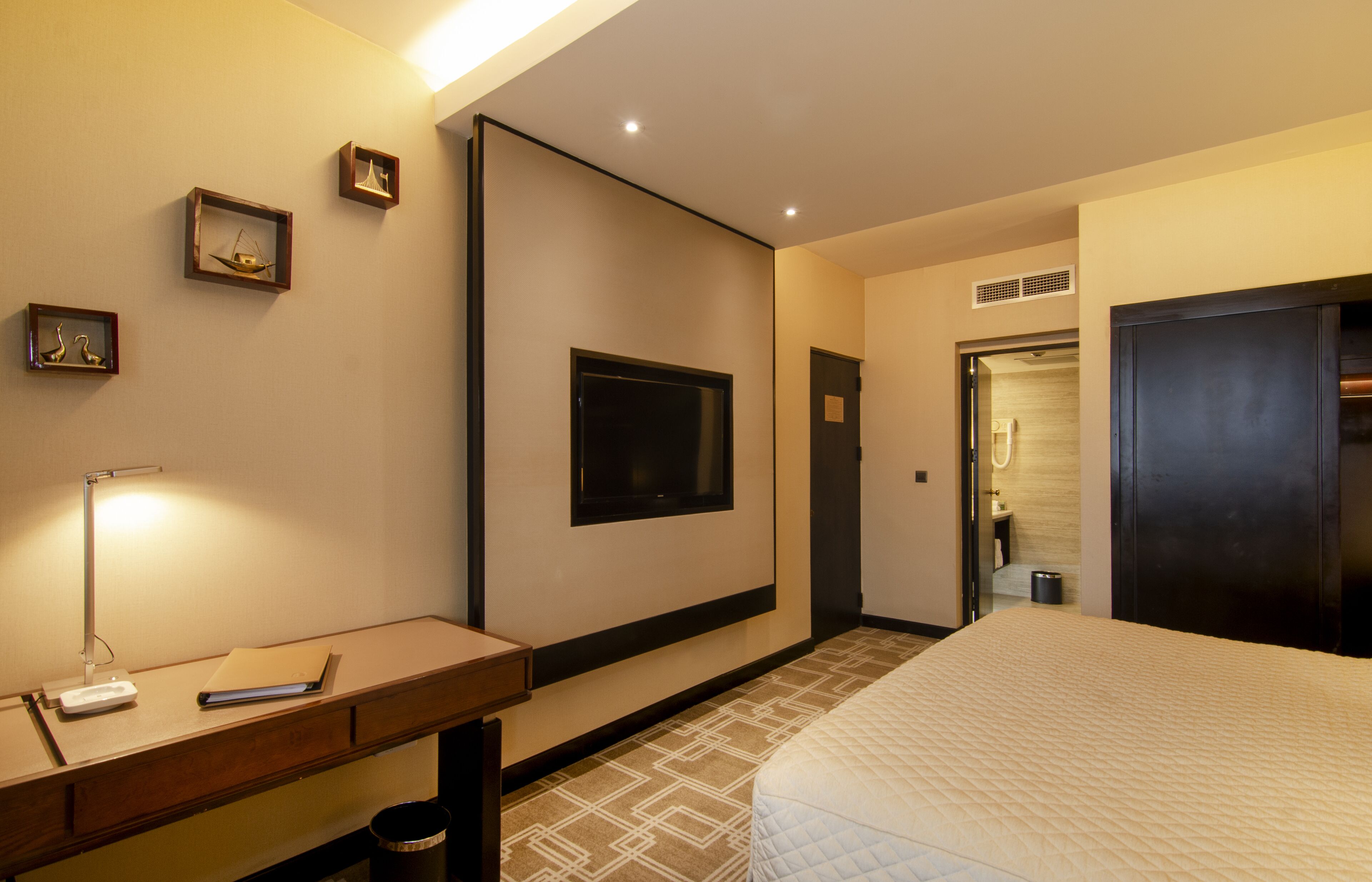 A hotel room with a bed, a desk with a lamp, a television, and framed pictures on the wall.