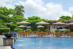 Uma área de piscina com espreguiçadeiras e guarda-sóis, rodeada por vegetação exuberante e palmeiras.