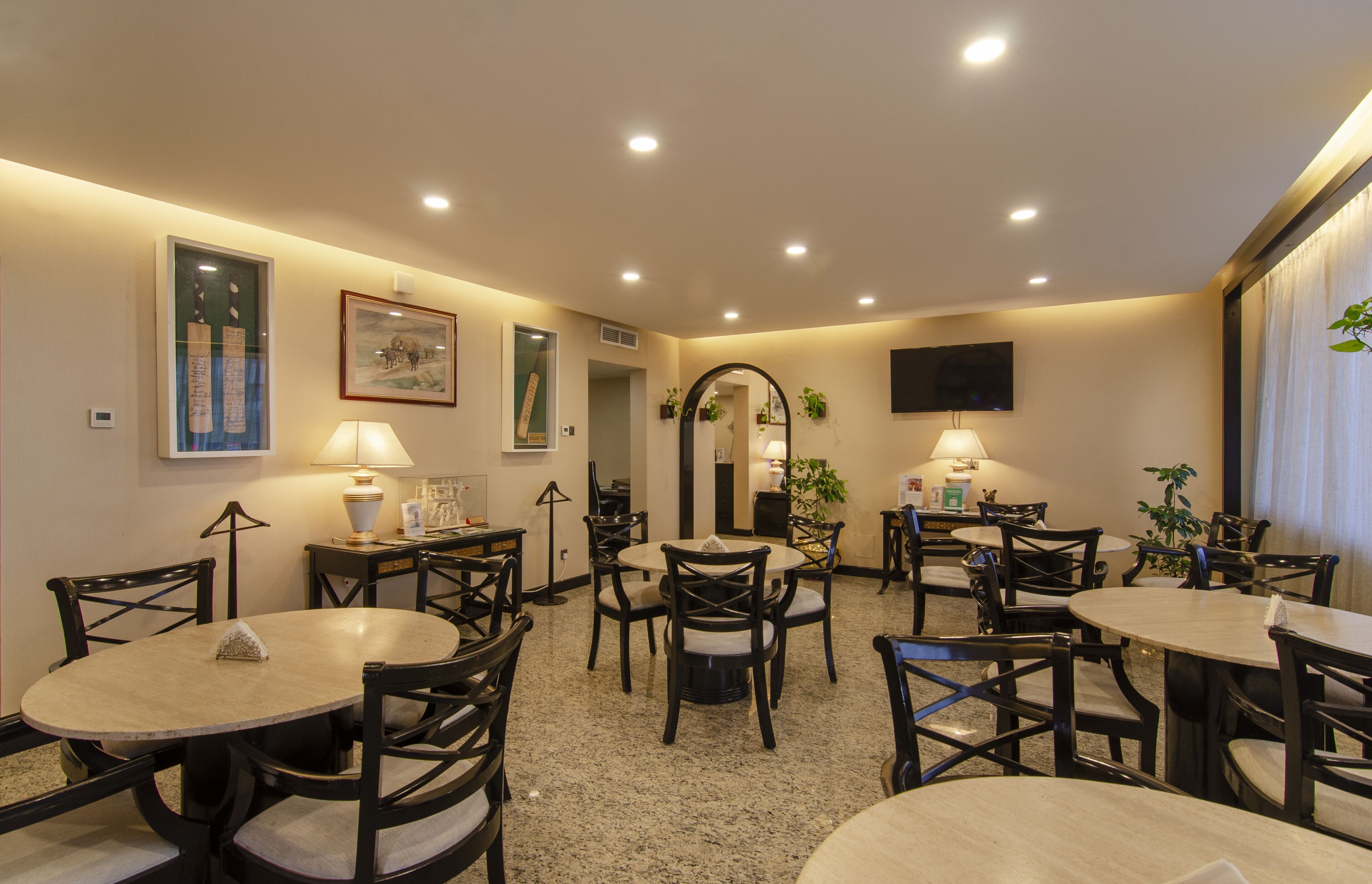 A dining area with round tables and chairs, recessed ceiling lights, framed artwork, and potted plants.