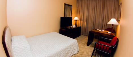 Quarto de hotel com cama, mesa de trabalho com cadeira, televisão e duas luminárias.