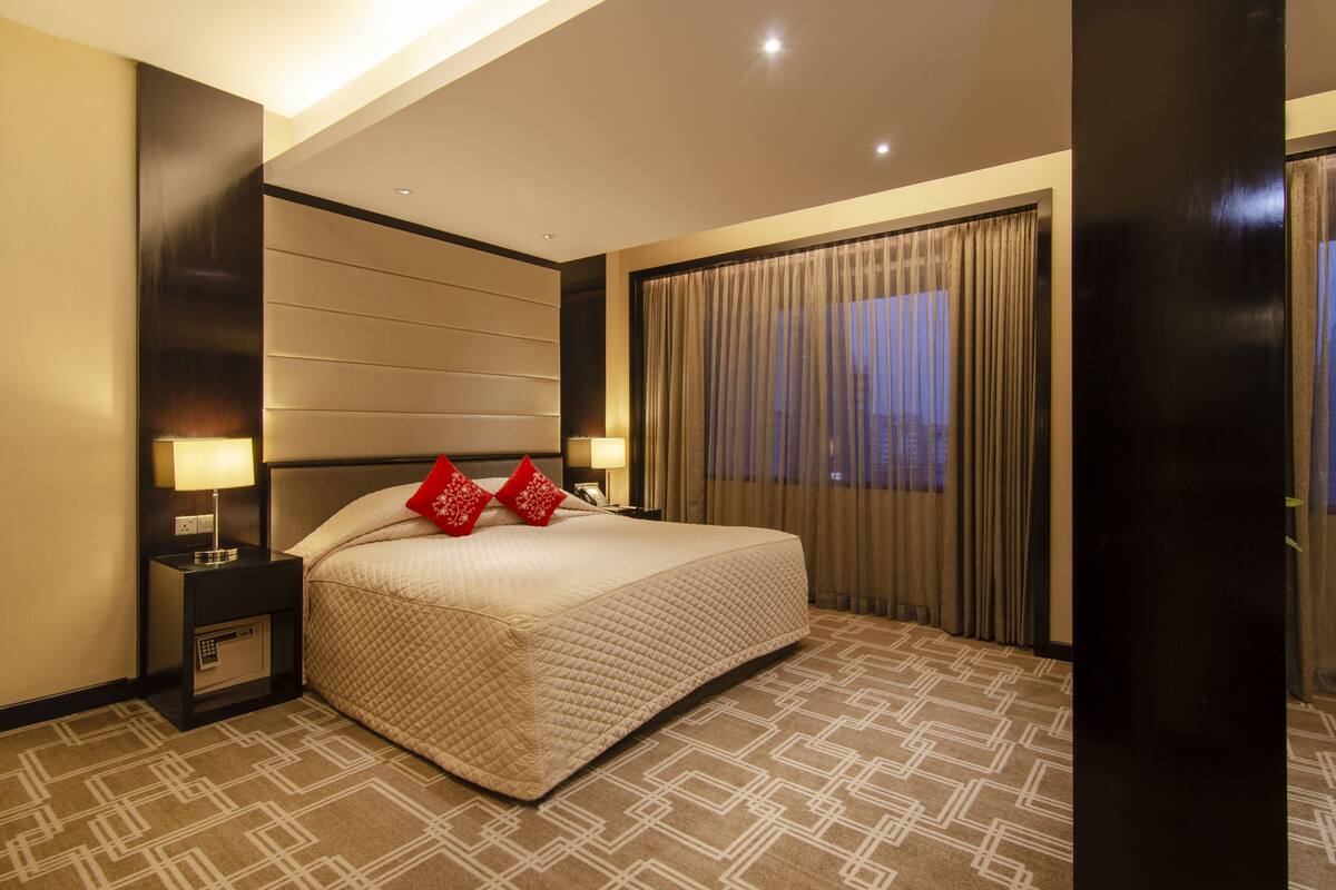 A hotel room with a large bed, two bedside tables with lamps, a window with curtains, and a patterned carpet.