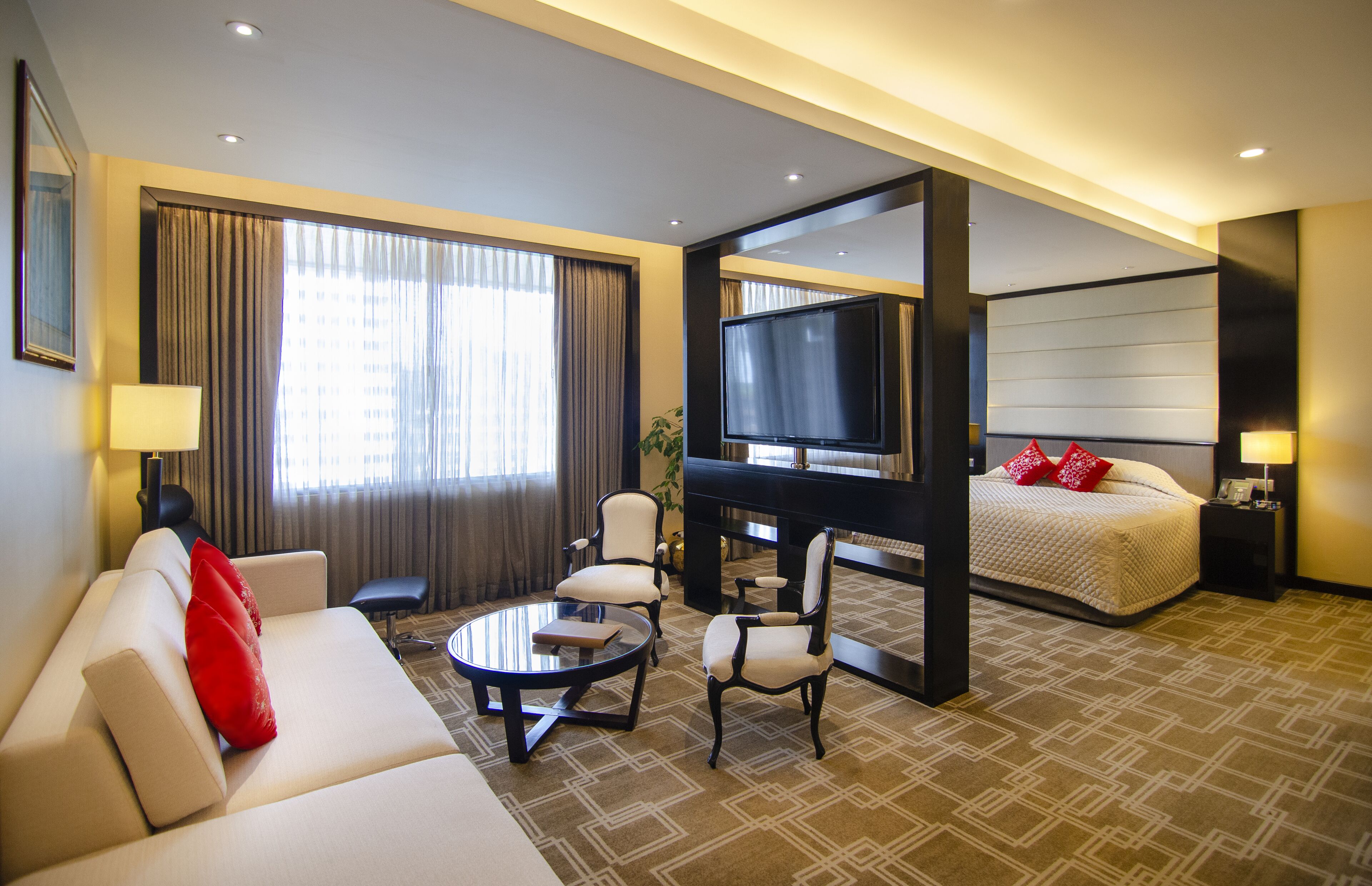 Camera d'hotel con divano, poltrone, tavolino, letto, televisore e scrivania con lampada.