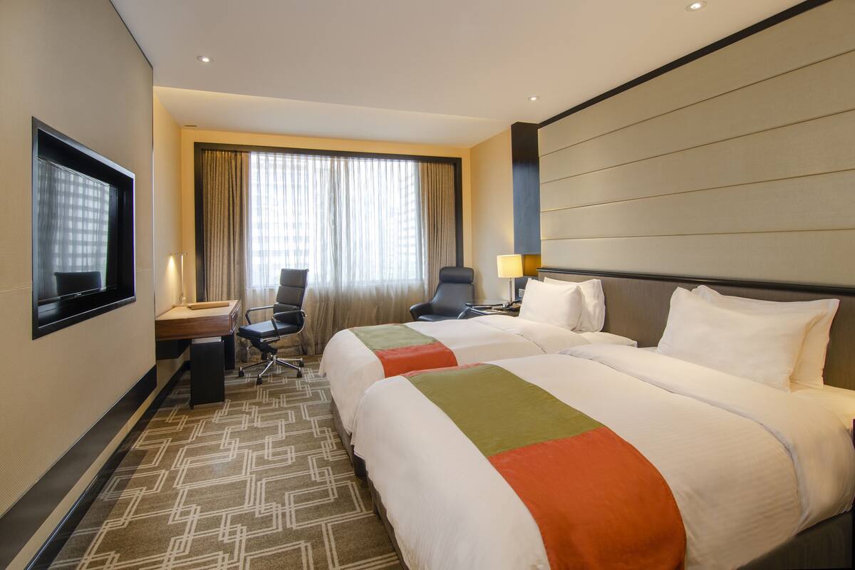 A hotel room with two beds, a desk, a chair, a TV, and a large window with curtains.