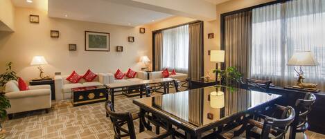Habitación de hotel con comedor, un conjunto de sofás y un amplio ventanal con cortinas.