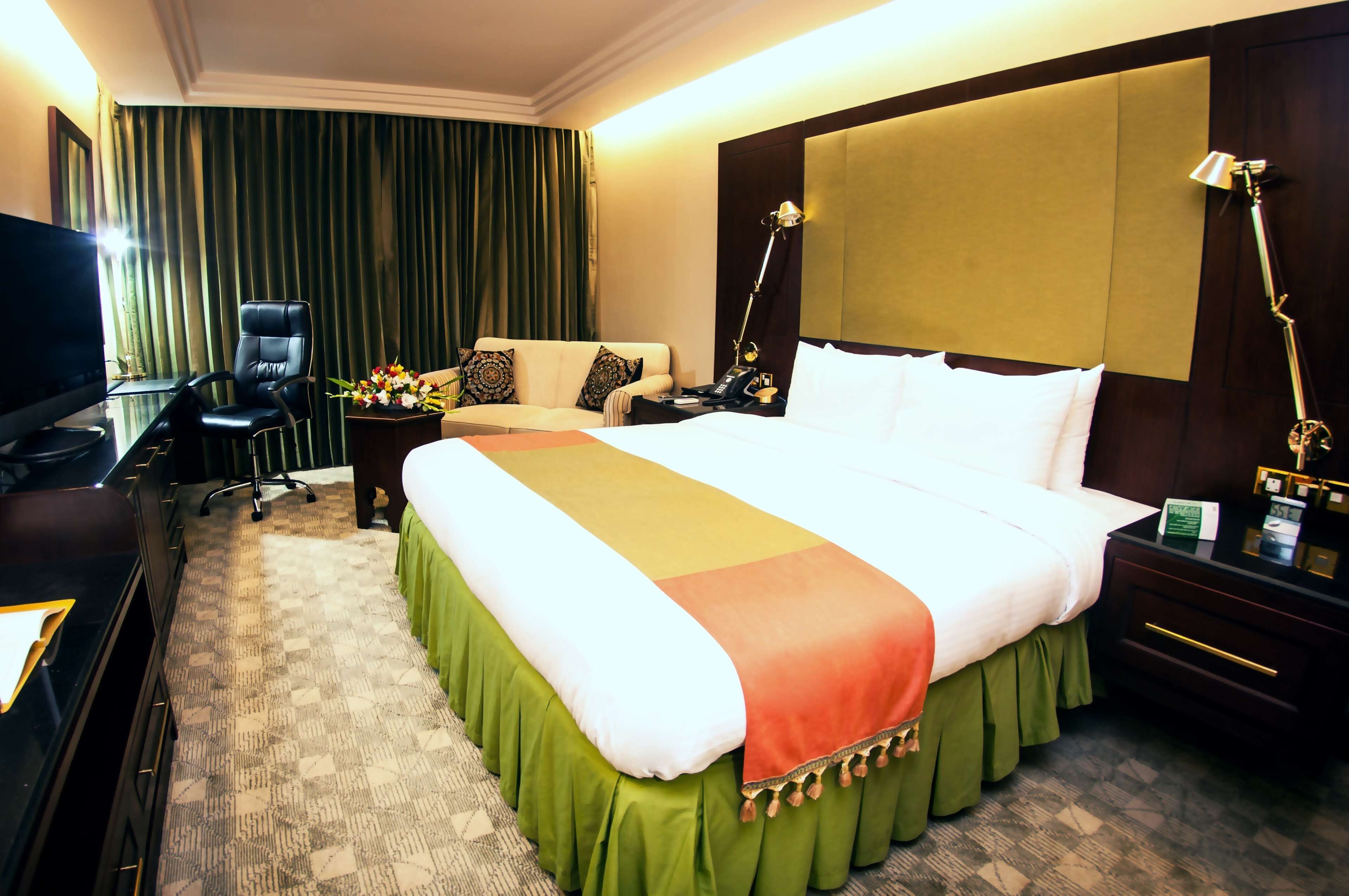 A hotel room with a large bed, a desk with a computer, a chair, a nightstand with a lamp, and a TV.